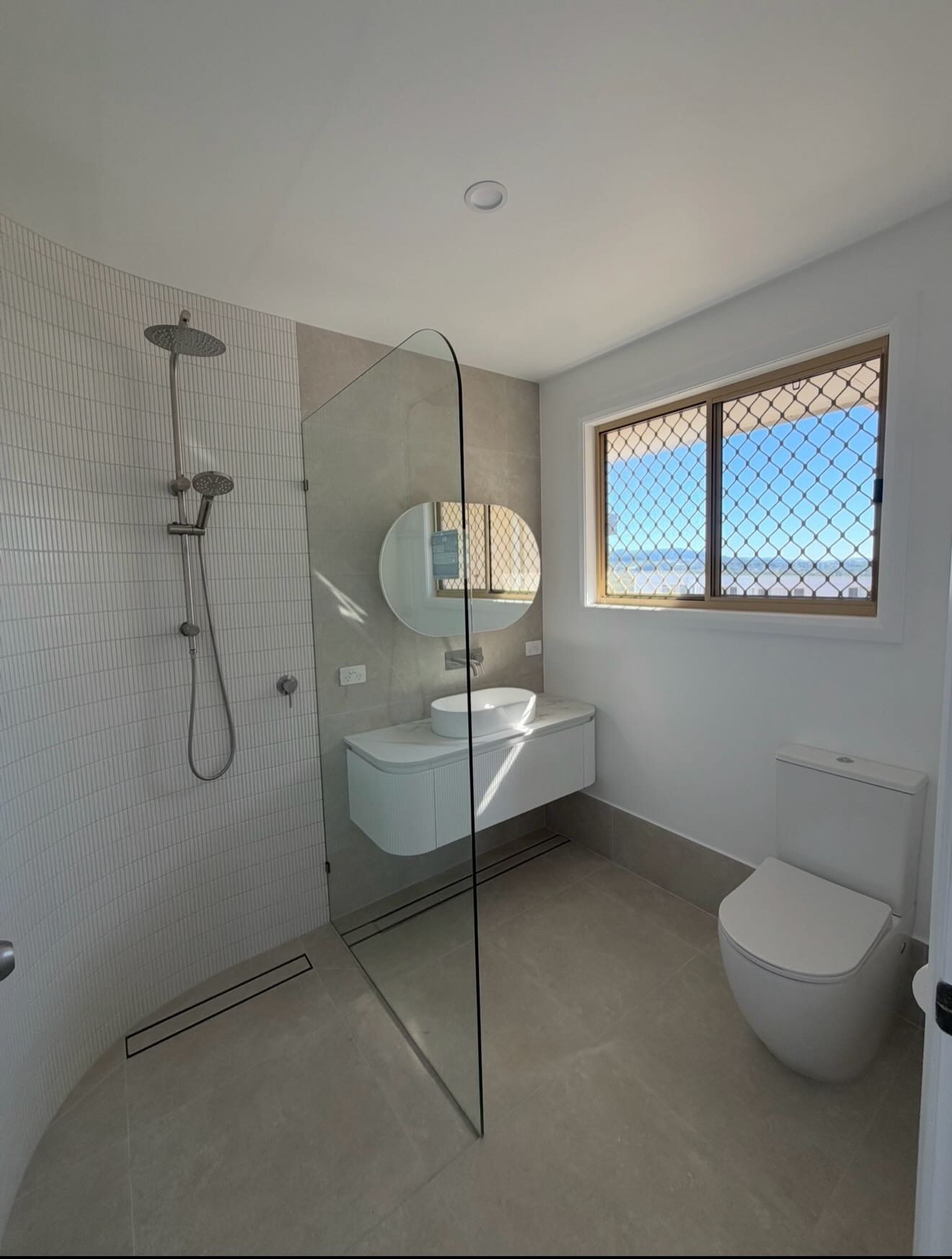 Completed bathroom renovation with curved tiled shower, oval mirror, floating vessel-sink vanity and a large window